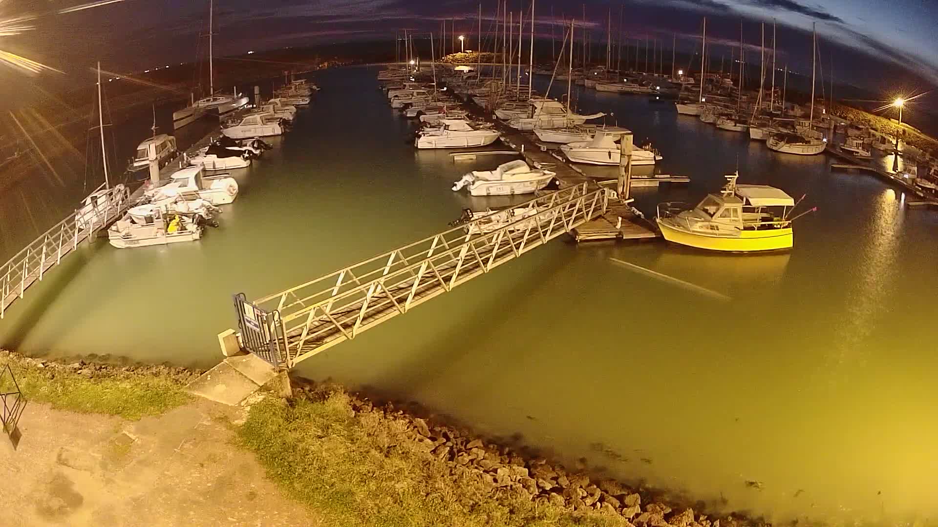 A marina at night, under a dark sky, with numerous boats docked alongside a metal walkway.