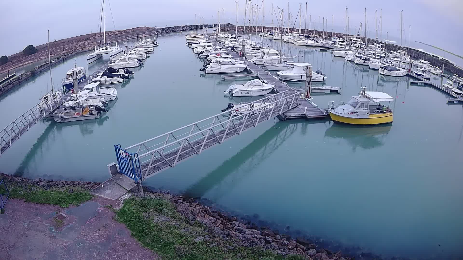 A marina filled with numerous boats and yachts on a calm, overcast day, connected by a metal walkway to the shore.