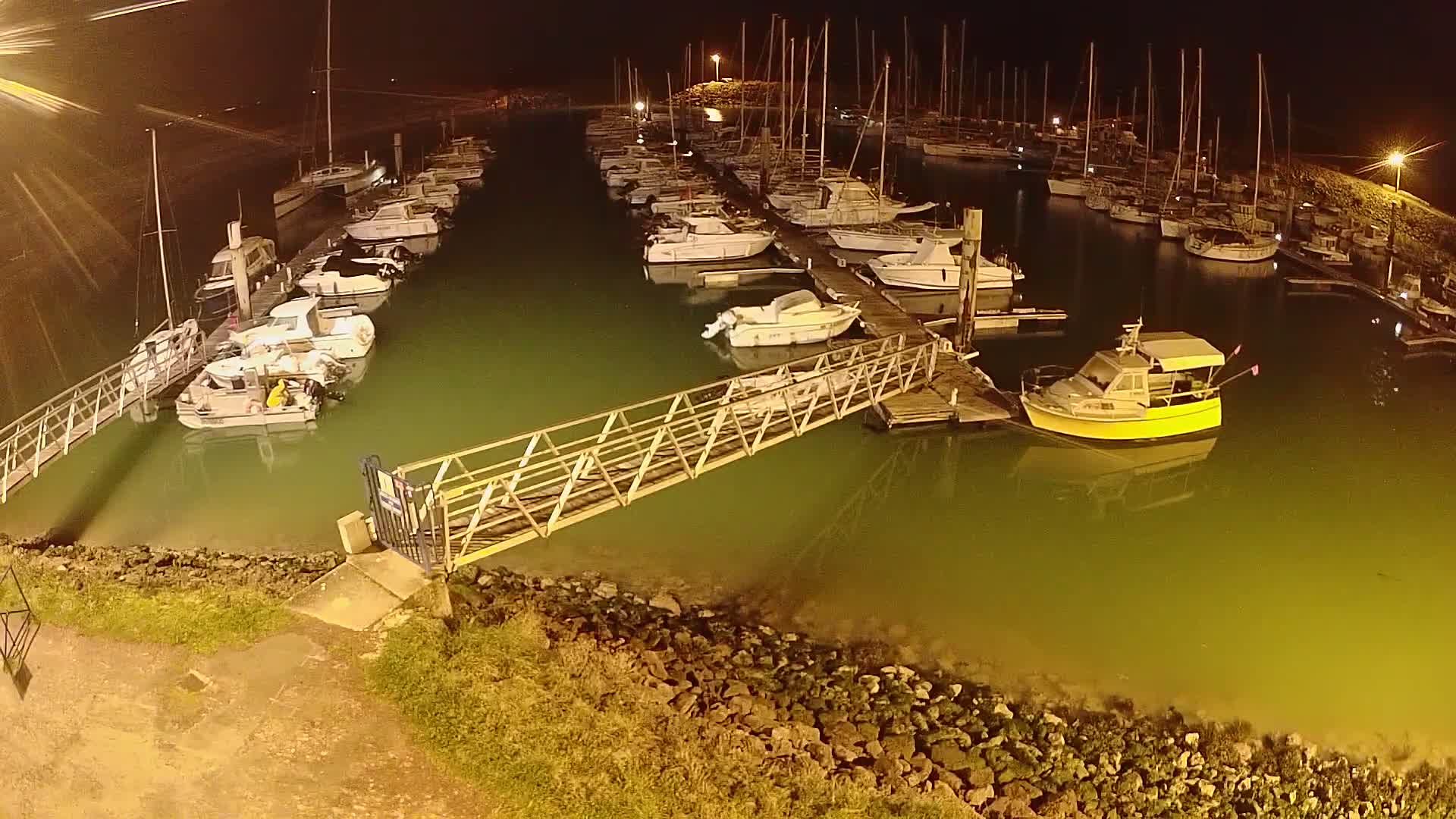 At night, a marina filled with numerous boats is illuminated, with a metal walkway extending from the shore to a dock.