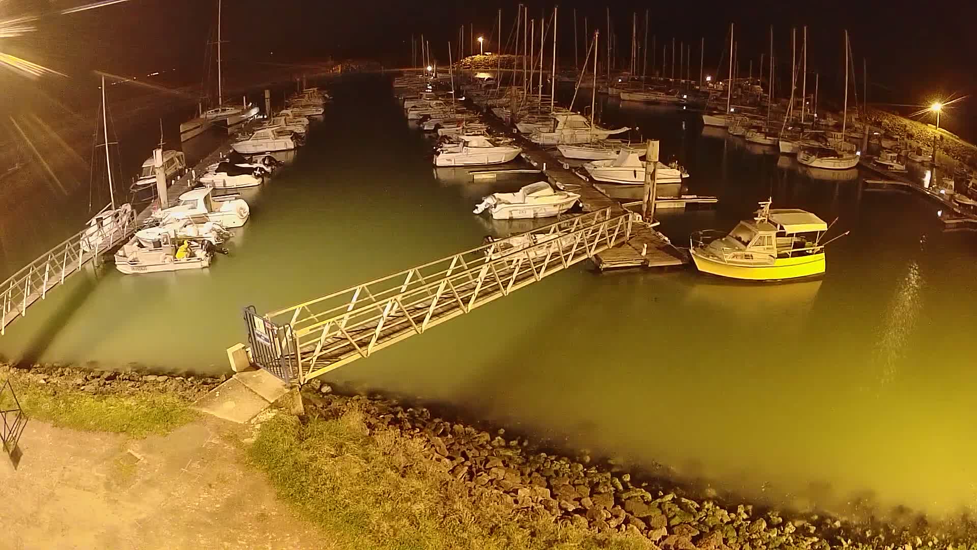 A marina at night, illuminated by artificial lights, shows numerous boats moored along docks, with a metal walkway extending from the shore to one of the docks.