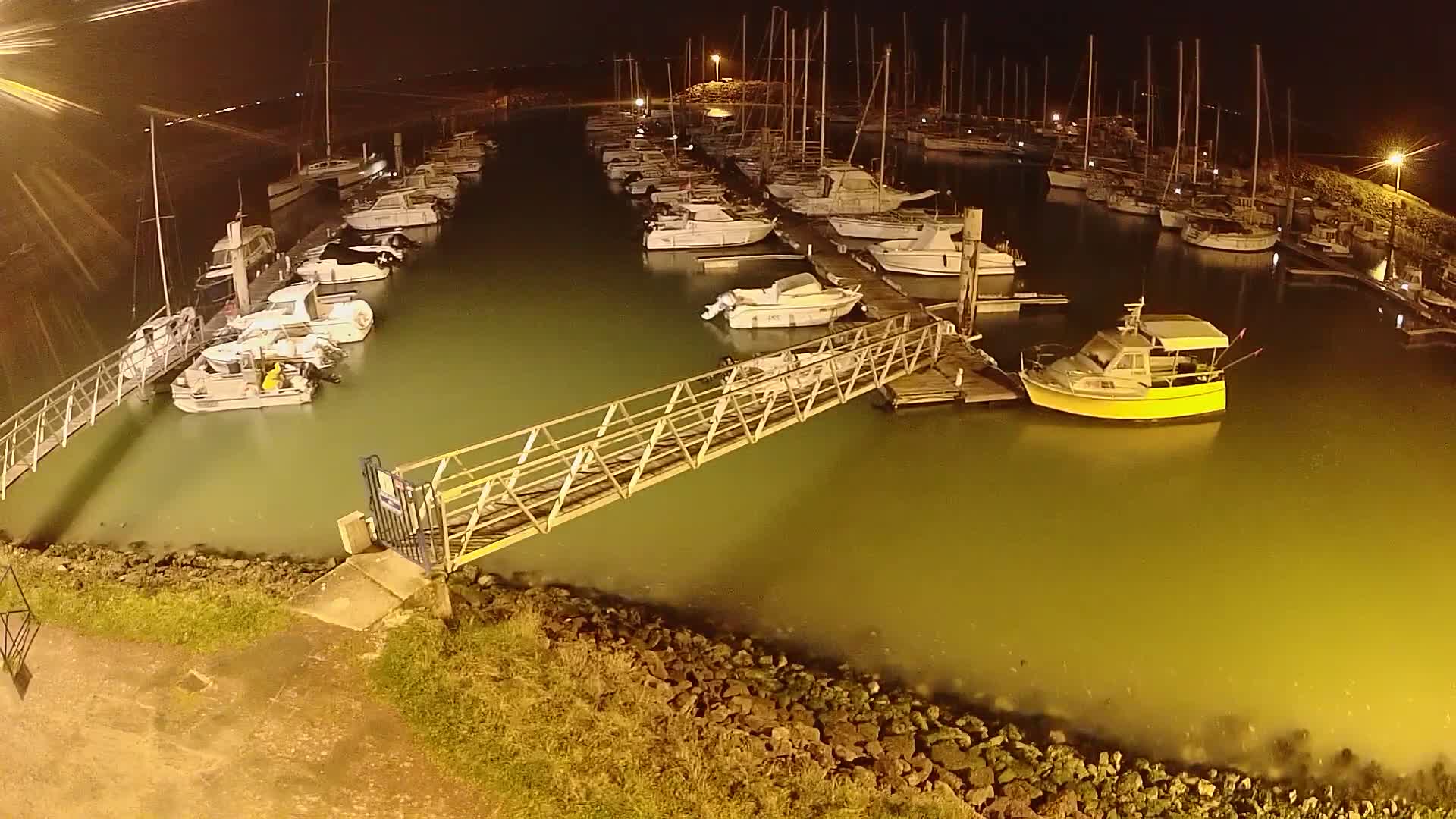 A marina at night, illuminated by artificial lights, shows numerous boats docked along piers, with a metal walkway connecting the shore to one of the docks.
