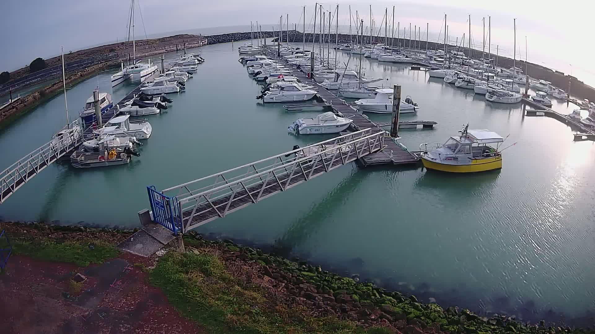 A calm marina with numerous sailboats and motorboats docked at piers, connected by a metal walkway, under a mostly clear sky.