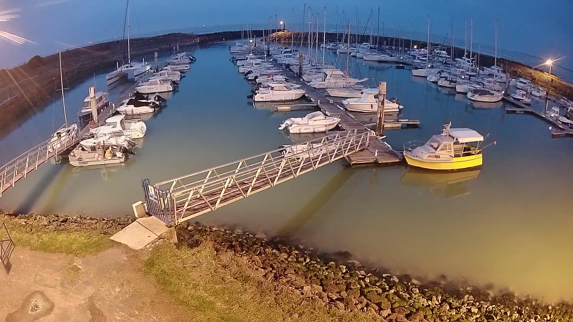 A marina at dusk, filled with numerous boats docked at piers, connected by a metal walkway.