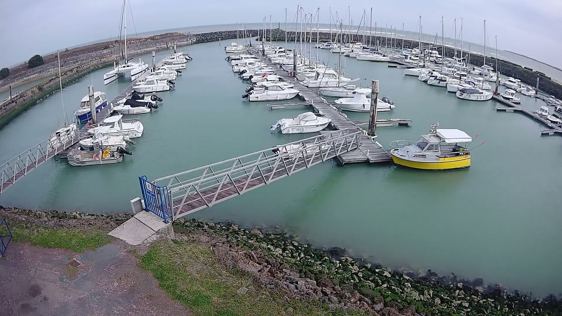 A marina filled with numerous boats on a cloudy day, connected by a metal footbridge to a rocky shoreline.