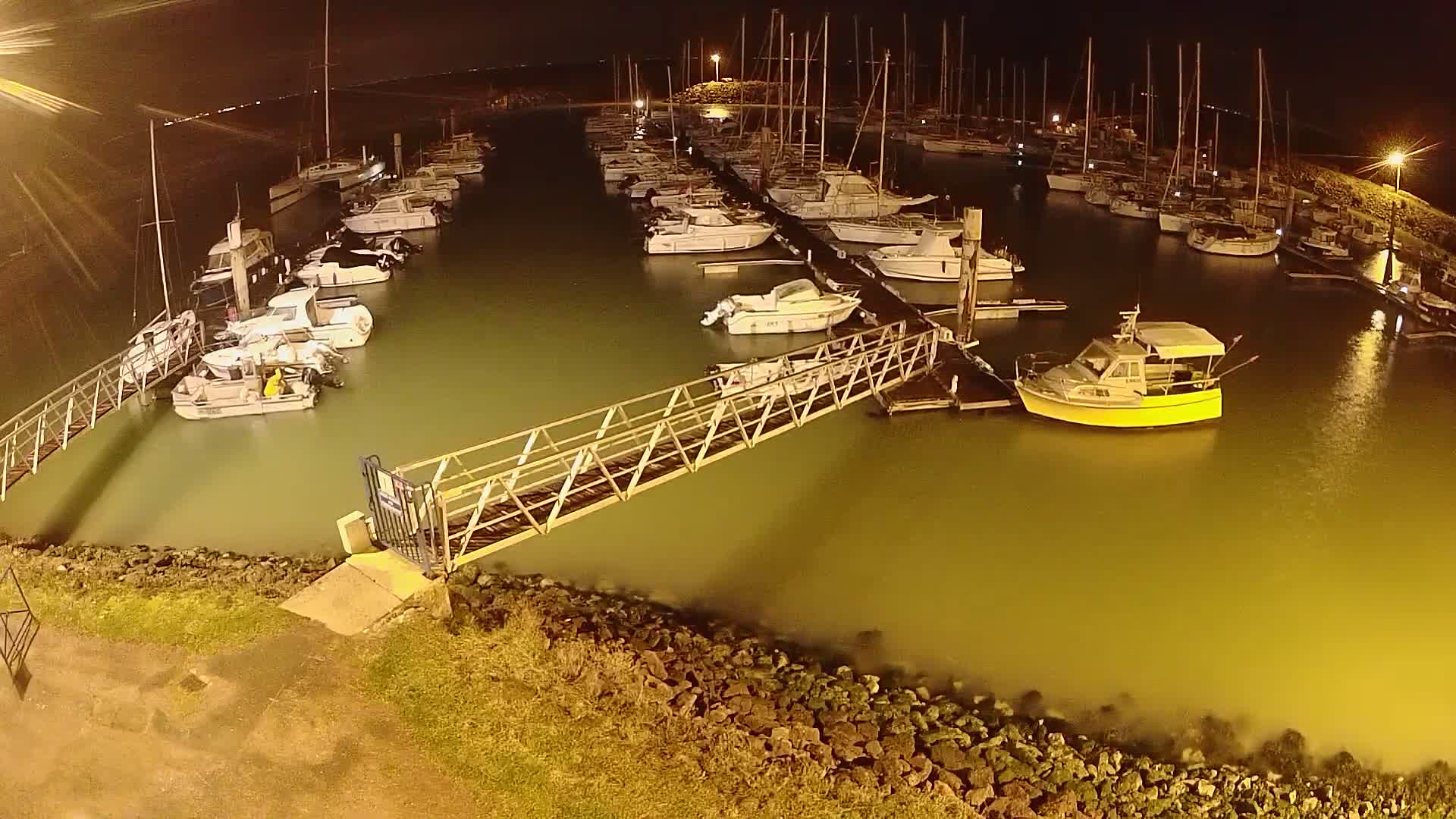 A metal walkway leads to a marina filled with numerous boats at night, under calm, dark conditions.