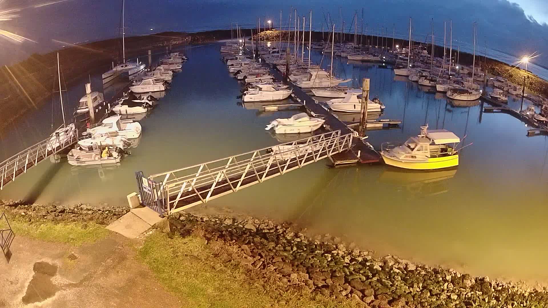 A marina at night, filled with numerous boats docked along a pier under a twilight sky.