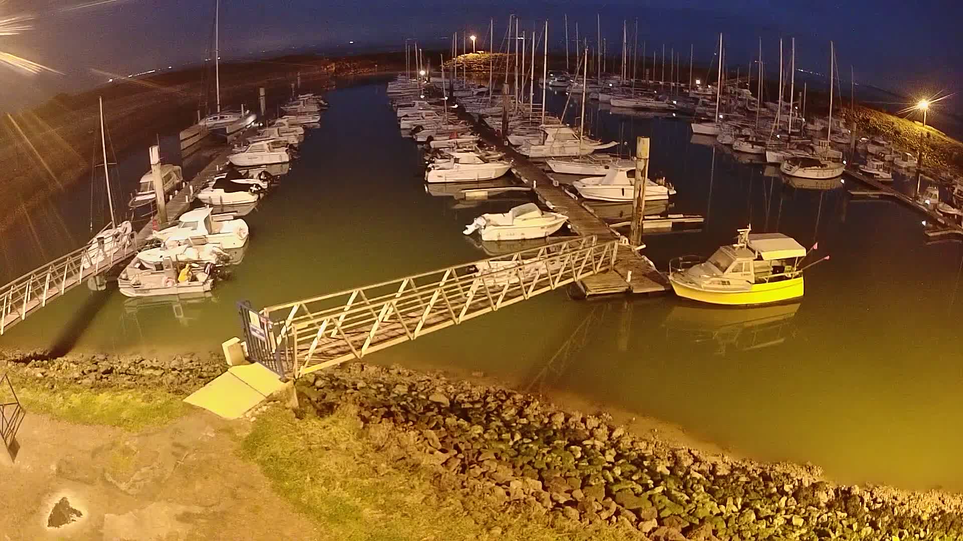 A marina at night, filled with numerous boats docked along piers, illuminated by artificial lights, under a dark sky.