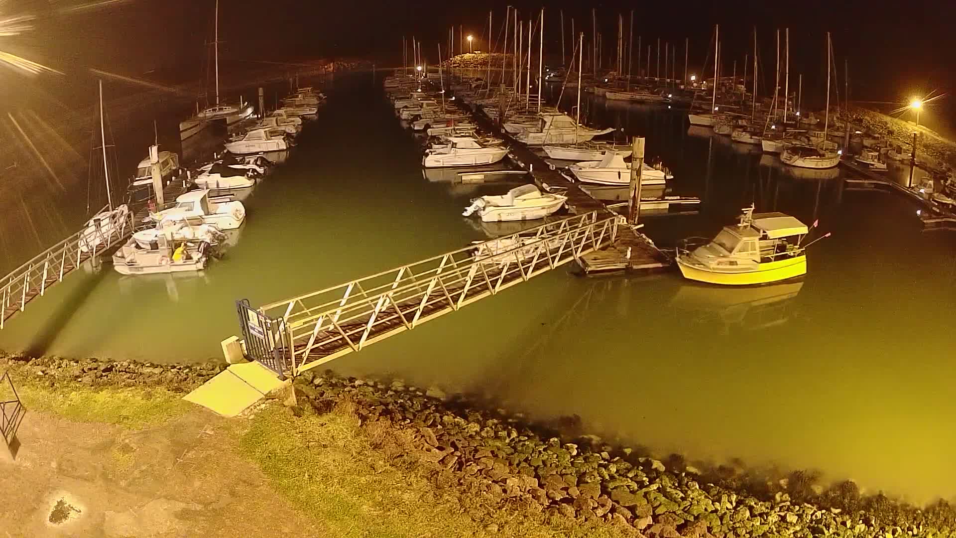 A marina at night, illuminated by artificial lights, shows many boats moored at docks, with a metal walkway leading to a small yellow boat.