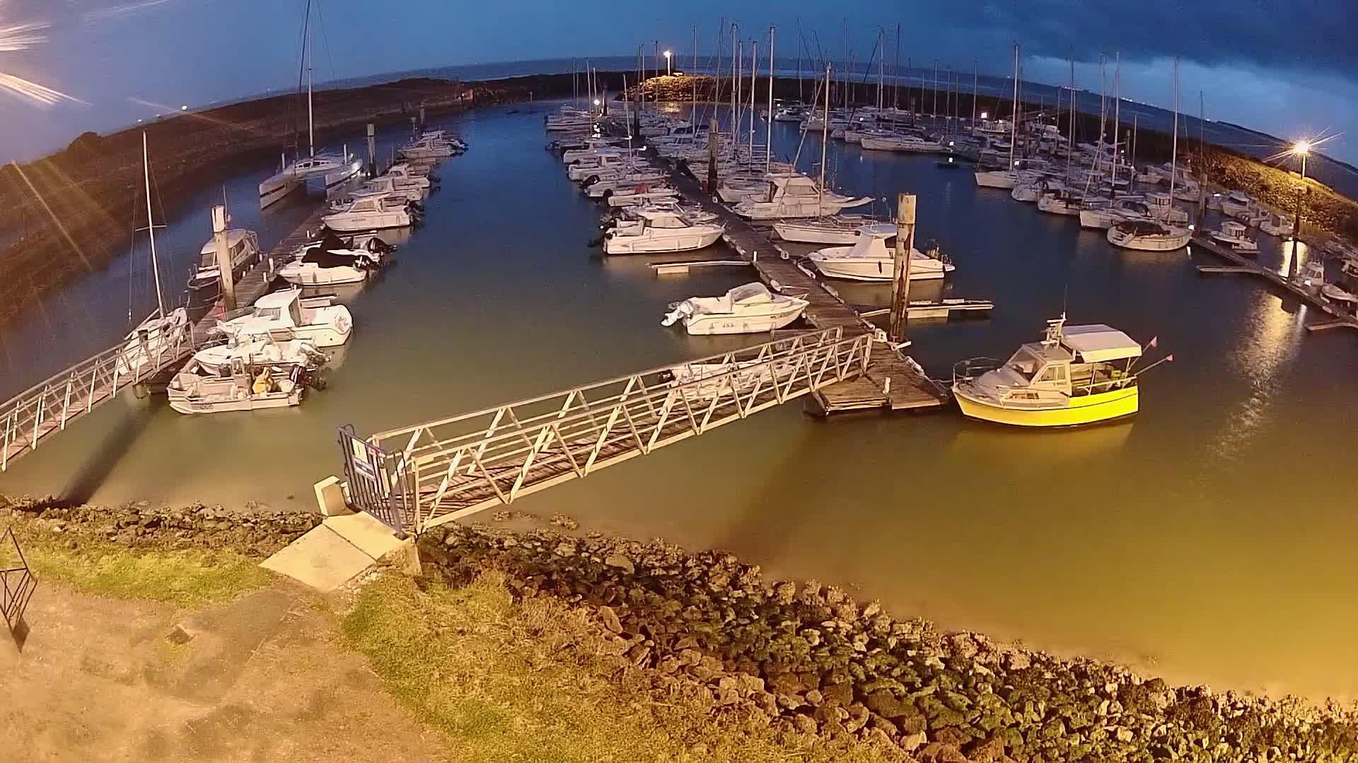 A marina at night, under a dark sky, is filled with numerous boats moored at docks, connected by a metal walkway.