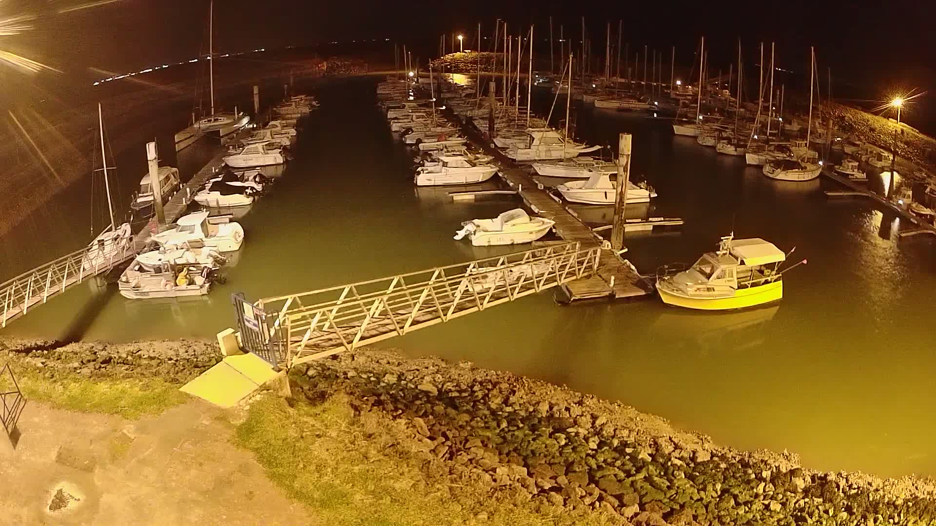 A marina at night, filled with numerous sailboats and motorboats docked along piers, is illuminated by artificial lights.