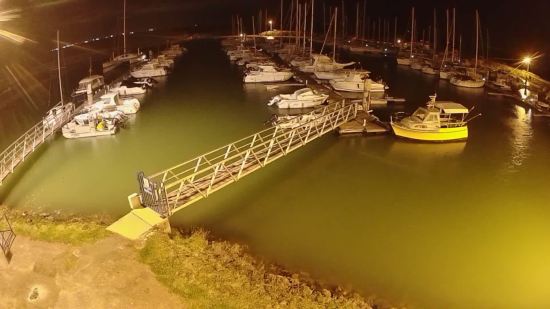 At night, a marina filled with boats is illuminated, with a metal walkway leading to a dock.