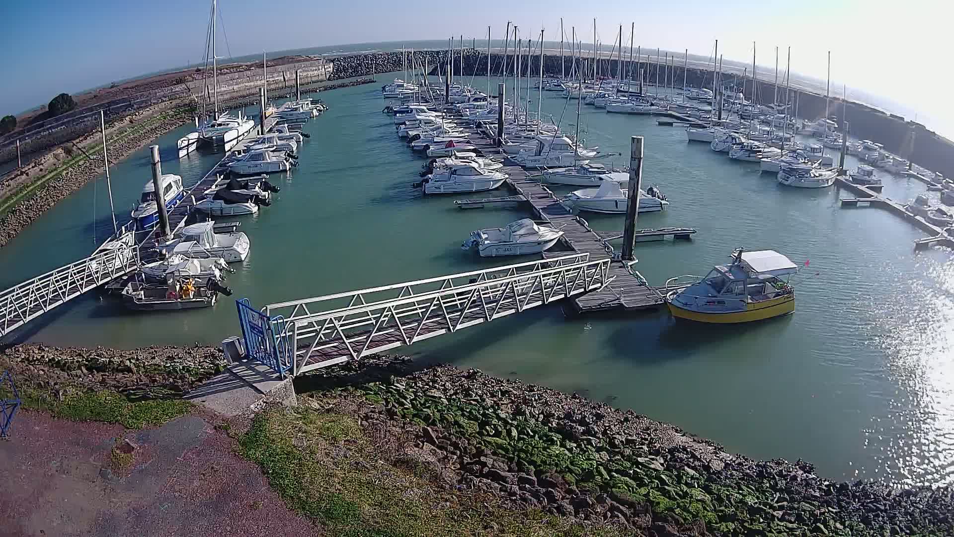 A marina filled with numerous sailboats and motorboats on a sunny day, with a metal walkway leading to a dock.