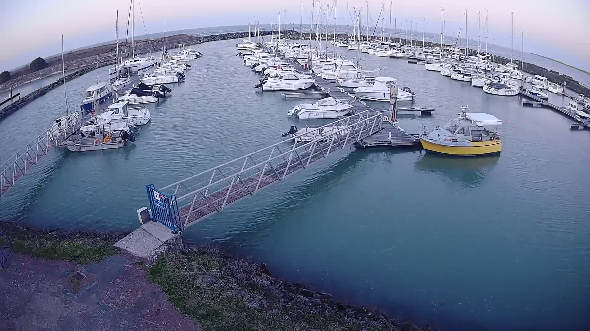 A marina filled with numerous sailboats and motorboats under a cloudy sky, connected by a metal walkway to the shore.