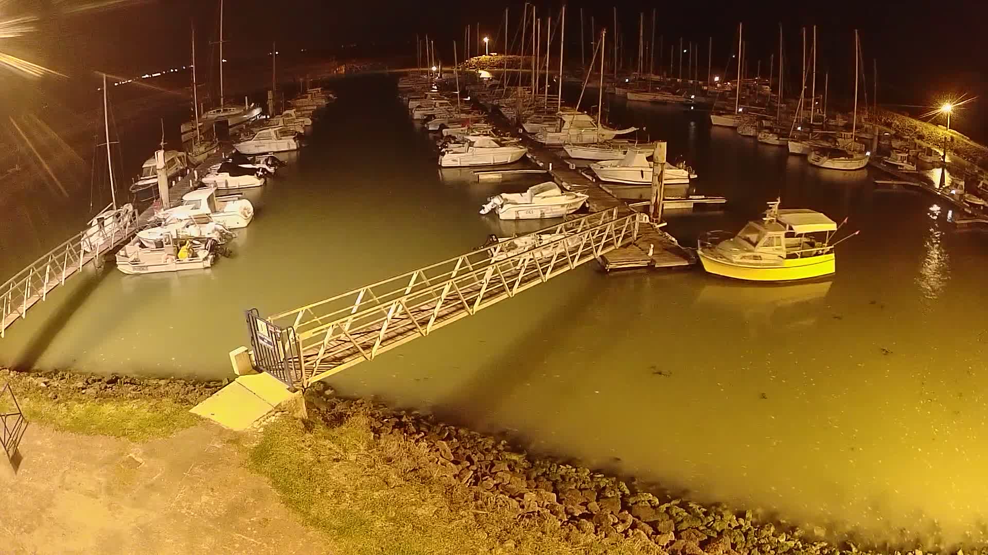 A marina at night, filled with numerous boats docked along piers and a yellow motorboat in the water, under somewhat murky, illuminated water.
