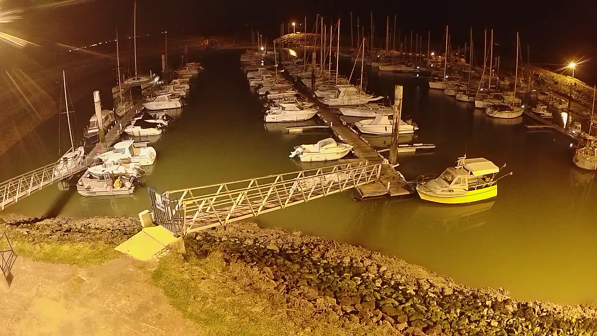 A marina at night, filled with numerous sailboats and motorboats docked along piers, is illuminated by artificial light.