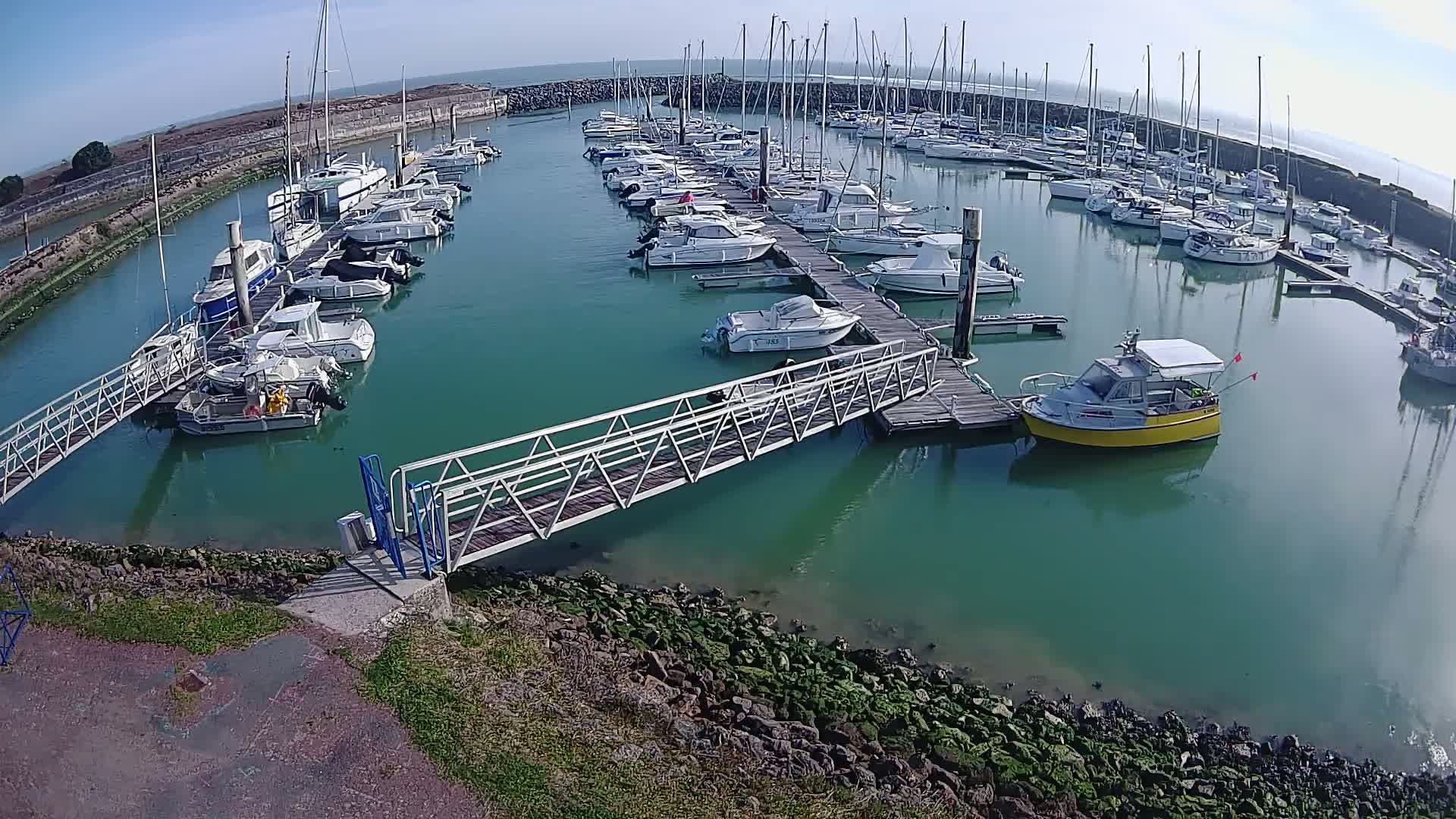 A marina filled with numerous sailboats and motorboats on a sunny day, with a metal walkway leading to a dock.