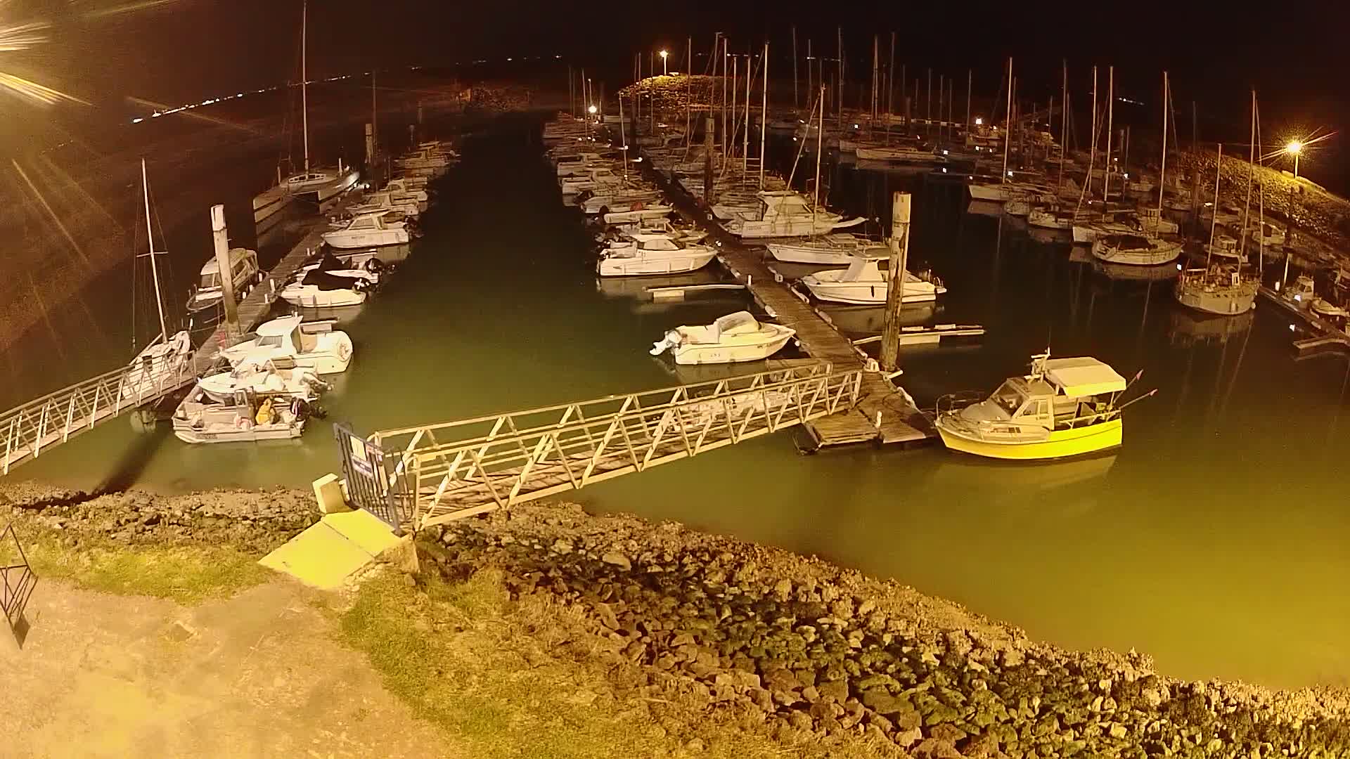 Many boats are docked at a marina at night, illuminated by artificial lights.
