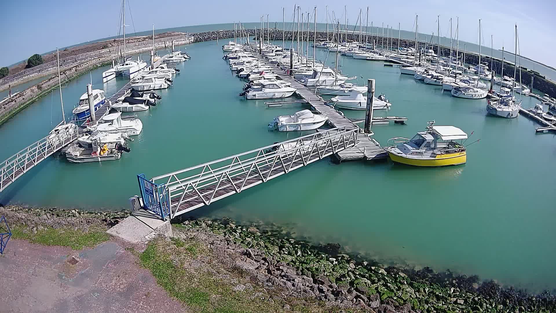 A marina filled with numerous sailboats and motorboats on a sunny day, with clear, teal-colored water and a metal walkway leading to a dock.