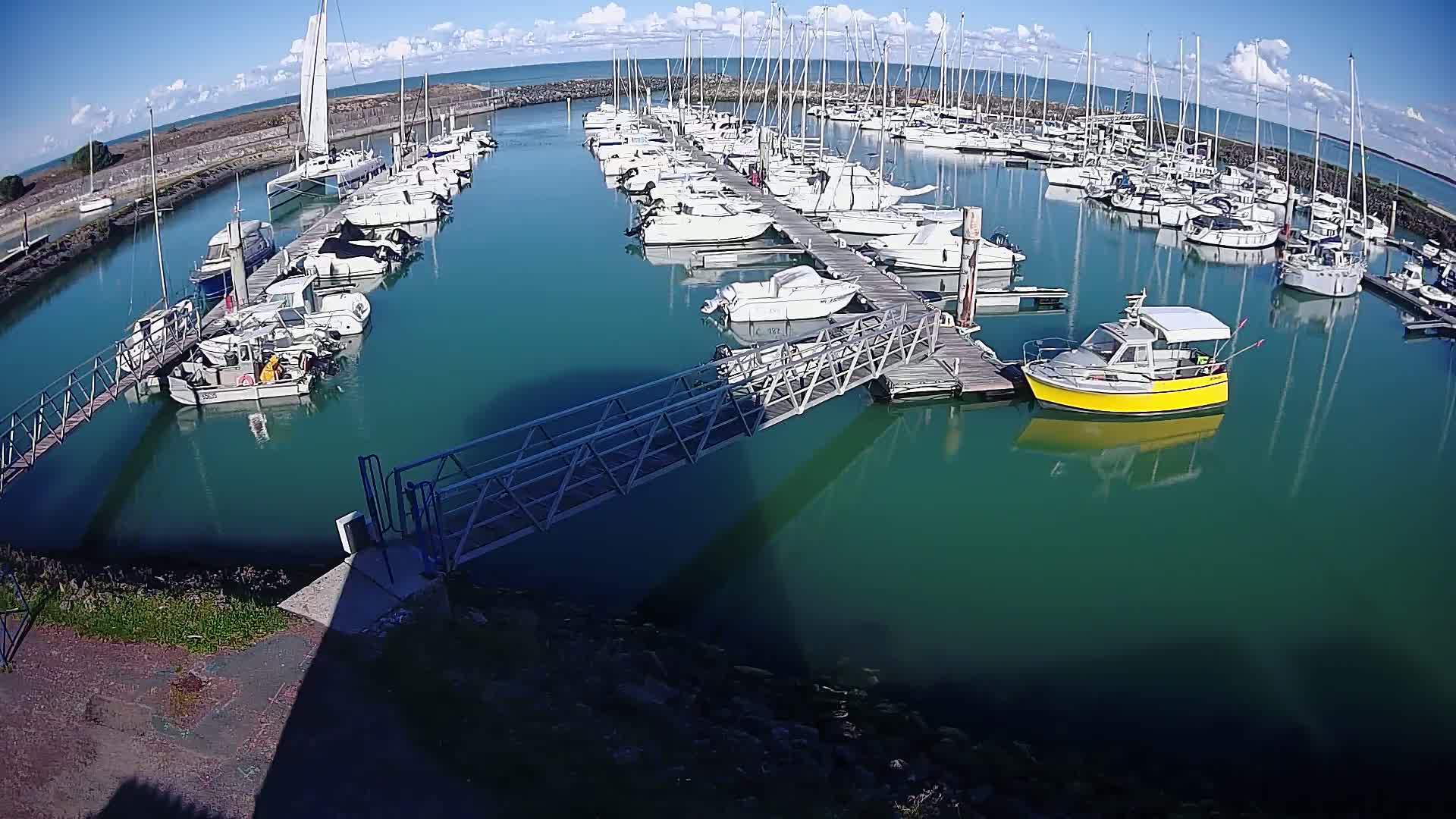 A marina filled with numerous sailboats and motorboats on a sunny day with partly cloudy skies and teal-colored water.
