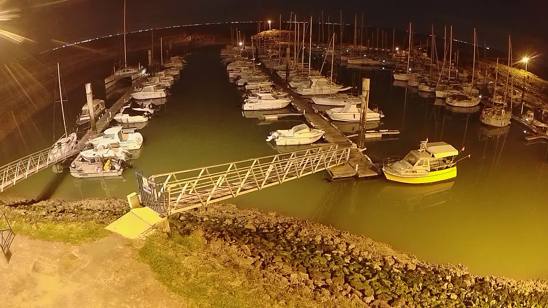 A marina at night, filled with numerous sailboats and motorboats docked along piers, illuminated by artificial light.