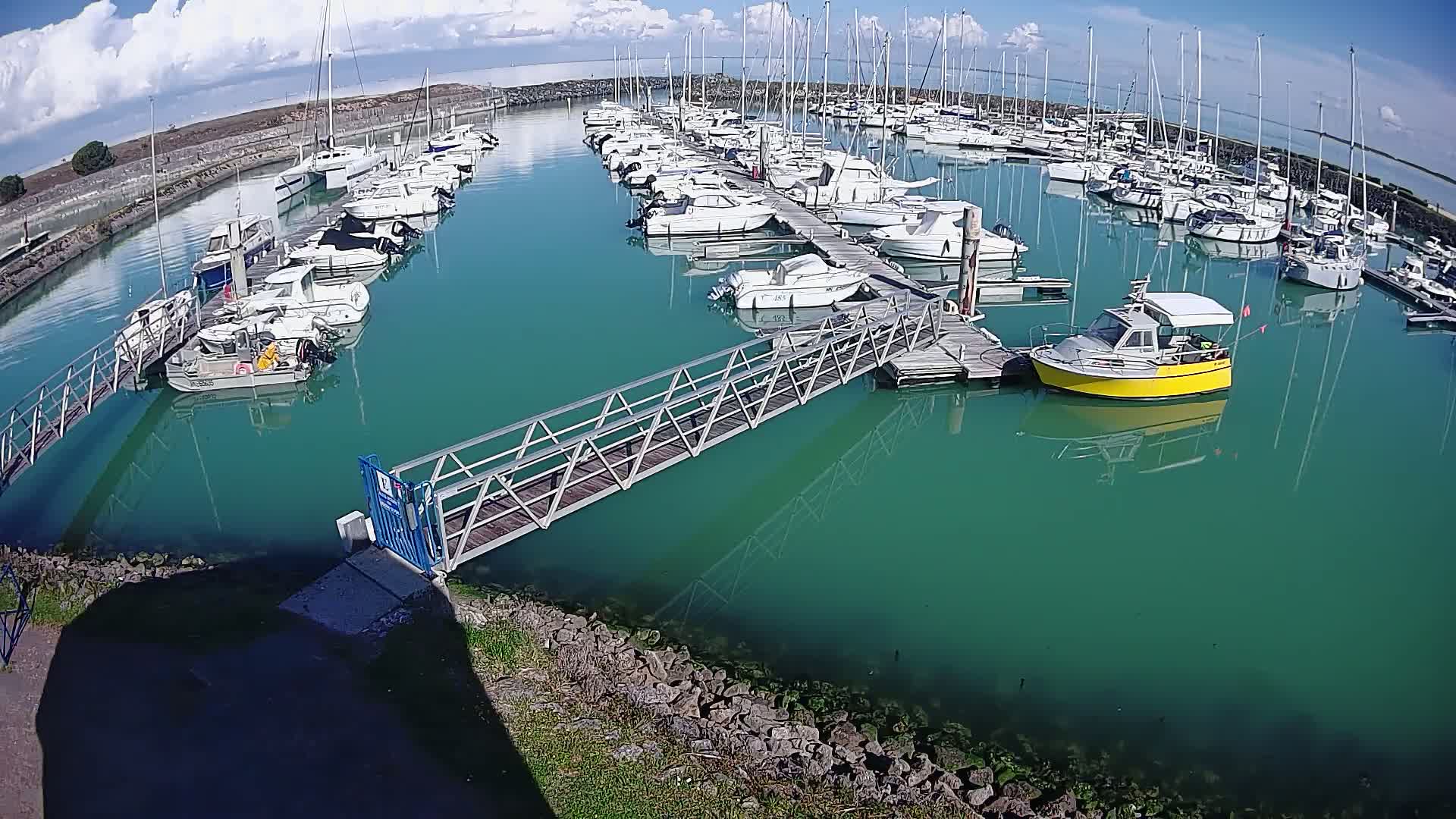 A marina filled with numerous sailboats and motorboats under a partly cloudy sky, with teal-colored water.
