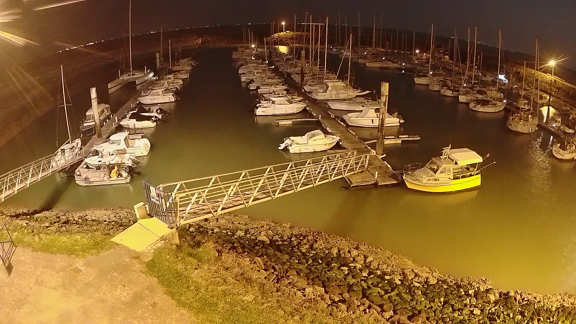 A marina at night, filled with numerous sailboats and motorboats docked along piers, illuminated by artificial light.