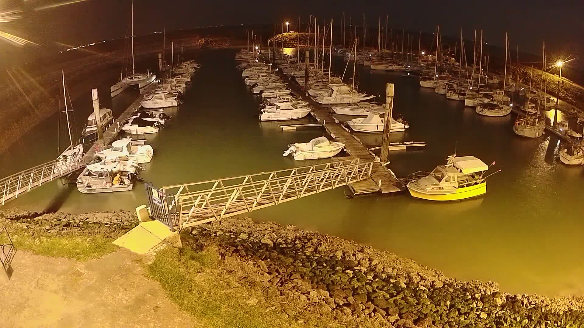 A marina at night, filled with numerous boats docked along piers, is illuminated by artificial lights.