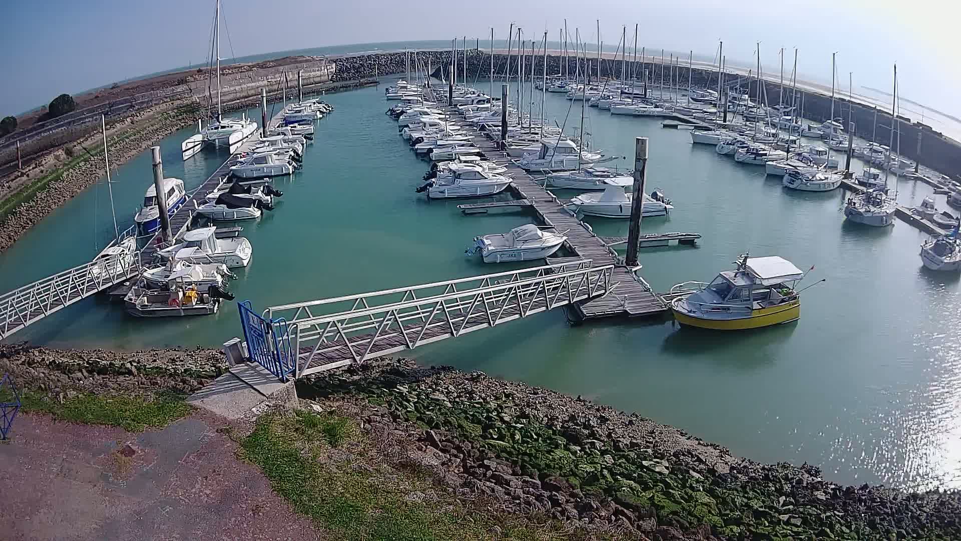 A marina filled with numerous boats on a sunny day, featuring a metal gangway leading to a dock.