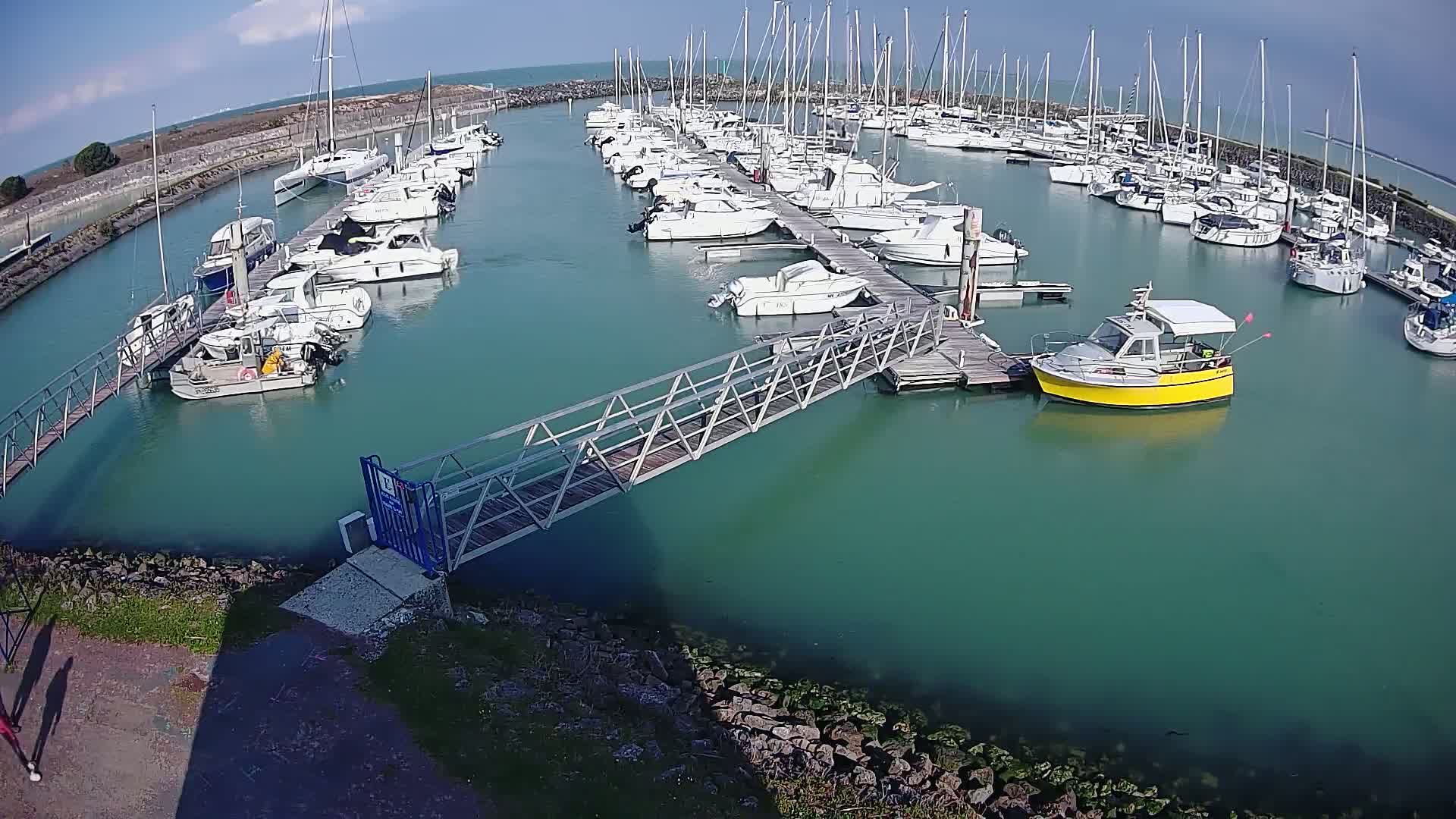 A marina filled with numerous sailboats and motorboats on a sunny day, connected by a metal walkway to the shore.