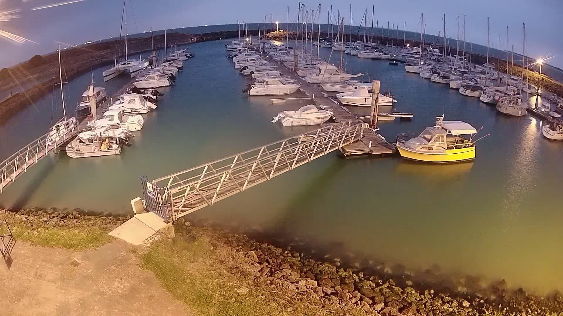 A marina at dusk, filled with numerous boats docked along piers, with a metal walkway extending from the shore to one of the docks.