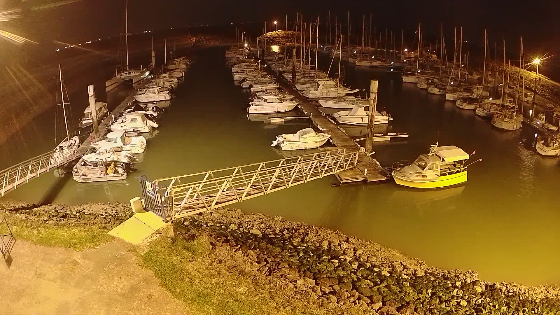 A marina at night, filled with numerous docked sailboats and motorboats, is illuminated under artificial light.