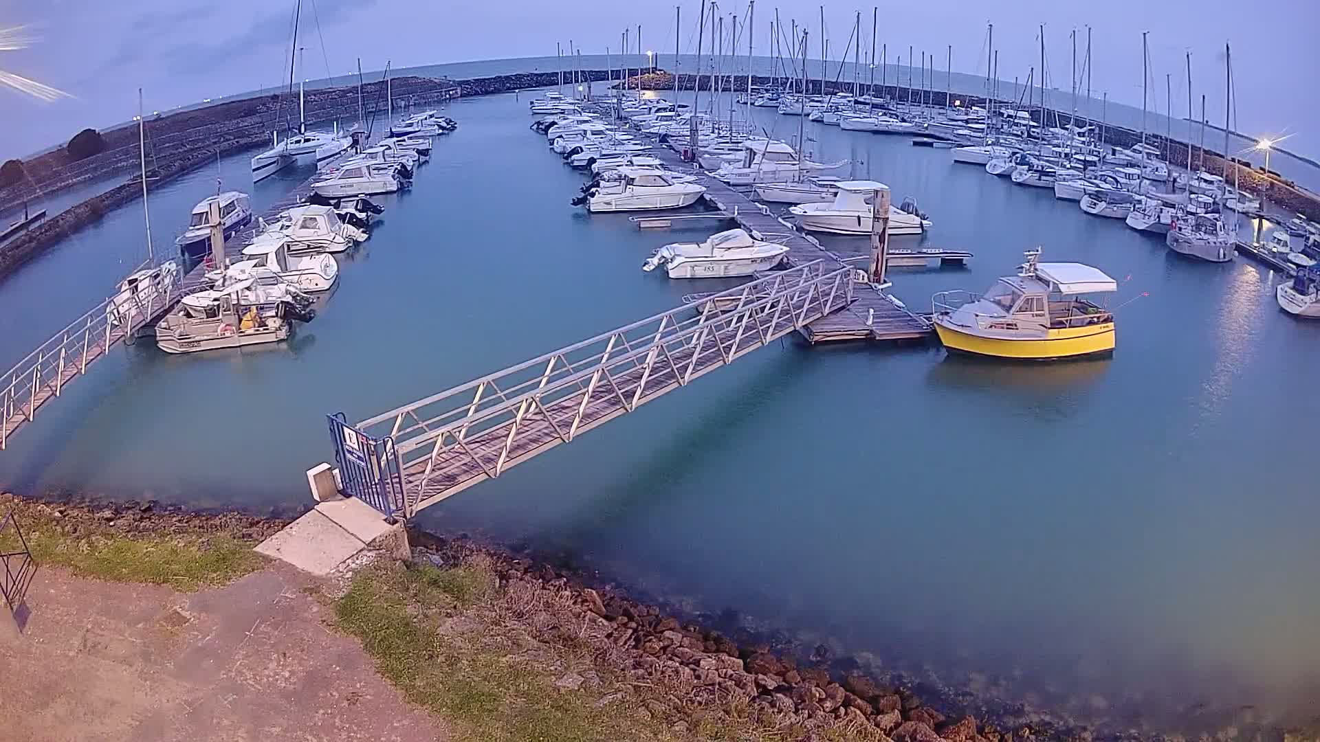 A marina filled with numerous boats of varying sizes is shown at dusk, with calm, teal-colored water.