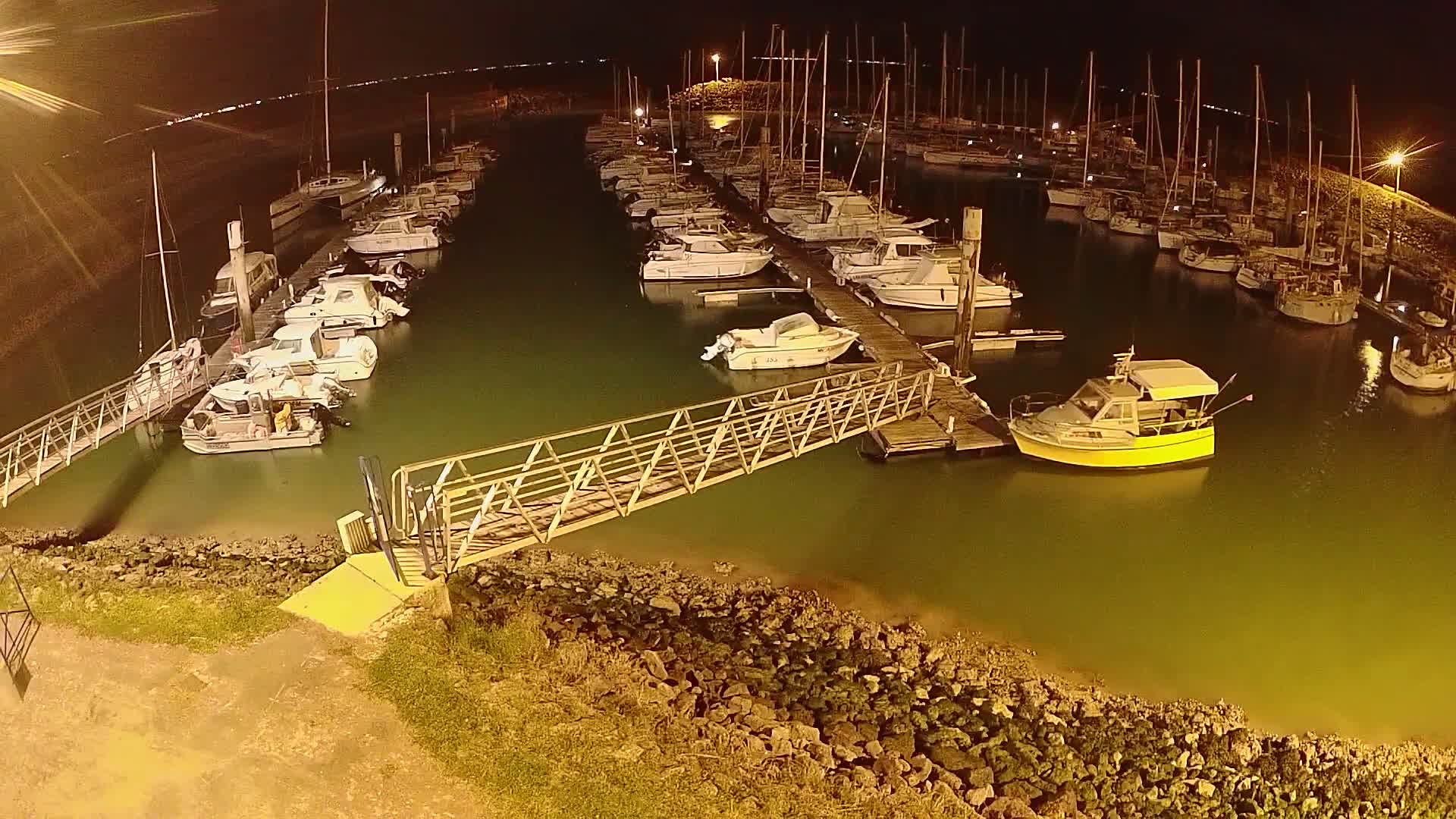 At night, a marina filled with numerous boats is illuminated, with a metal walkway extending from the shore to a dock.