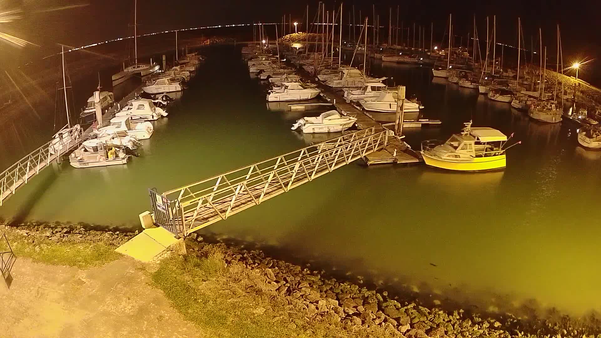 A marina at night, filled with numerous boats docked along piers, illuminated by artificial lights, with a metal walkway extending from the shore to one of the piers.