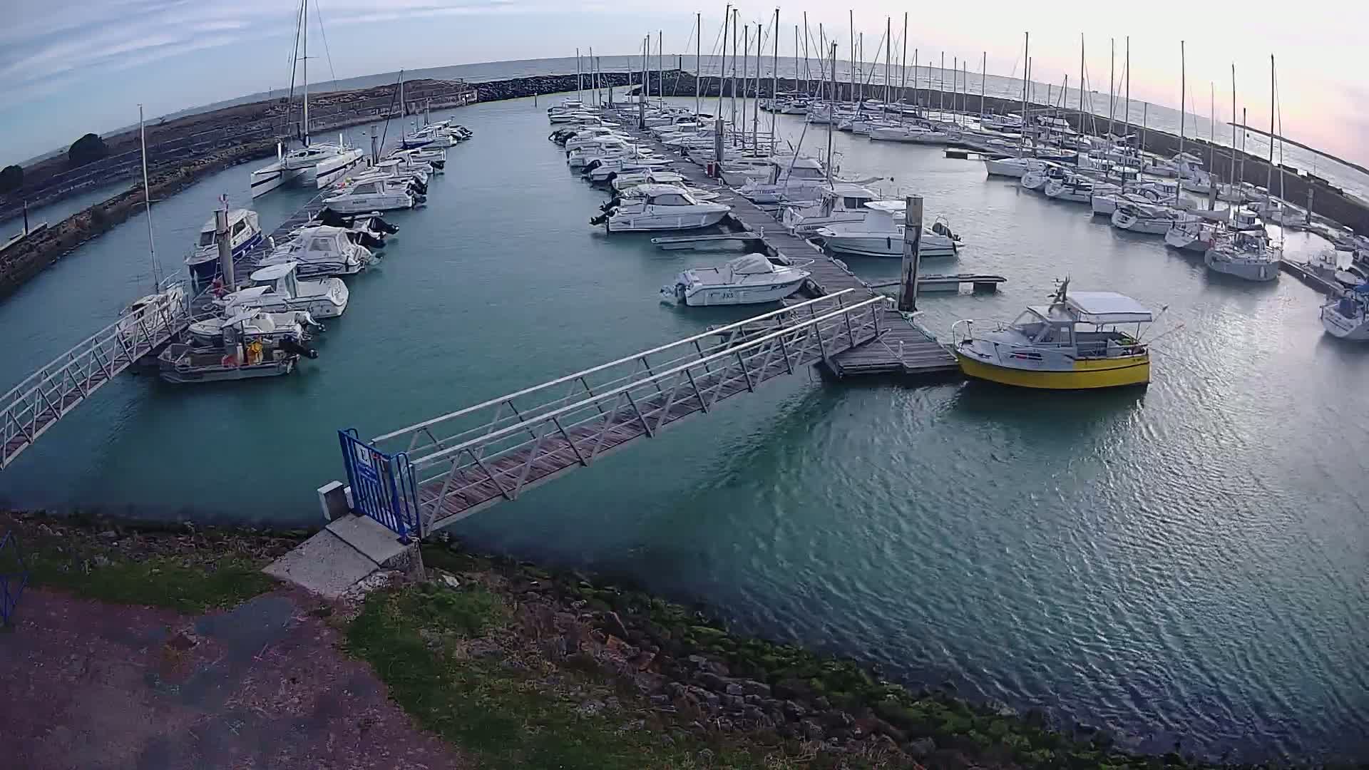 A marina filled with numerous sailboats and motorboats is calm under a partly cloudy sky.