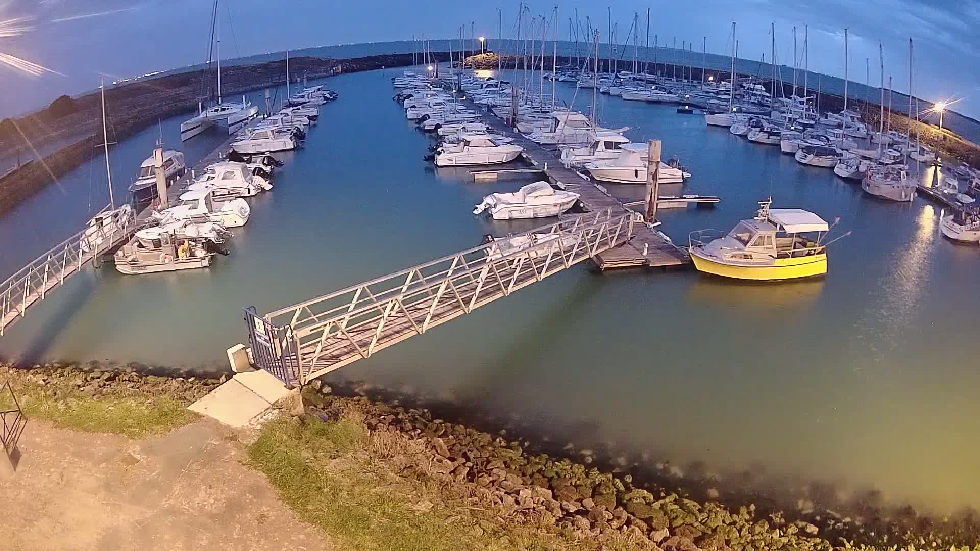 Many boats are docked at a marina at twilight, with a metal walkway leading from the shore to a pier.