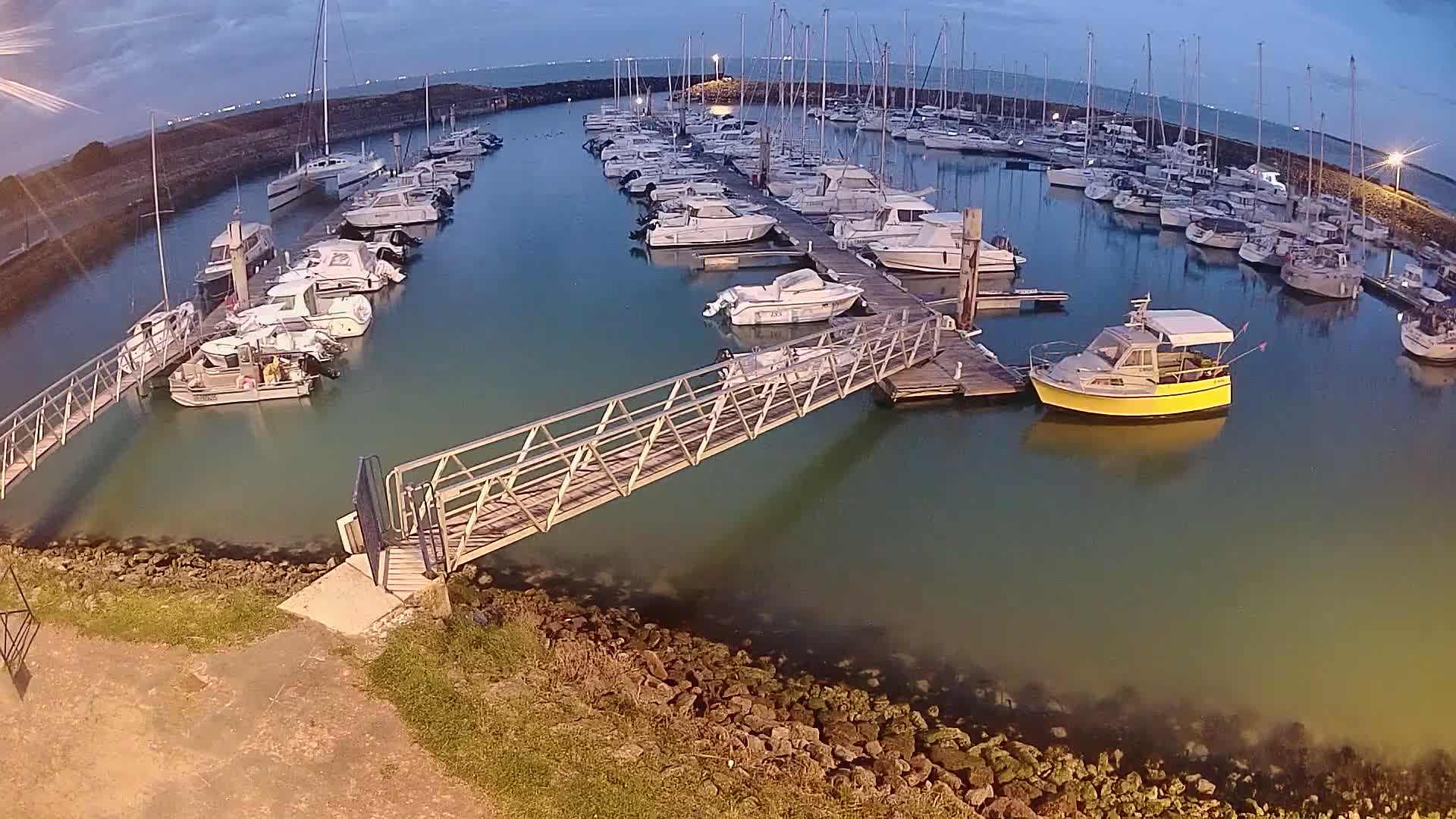 A marina filled with numerous boats at dusk, under a mostly clear sky, is connected by a metal walkway to the land.