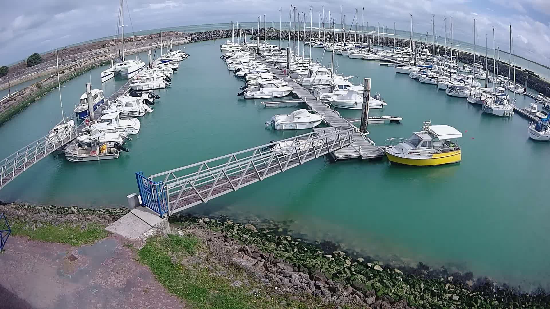 A marina filled with numerous boats of varying sizes is seen on a partly cloudy day, with a metal walkway leading from the shore to the docks.