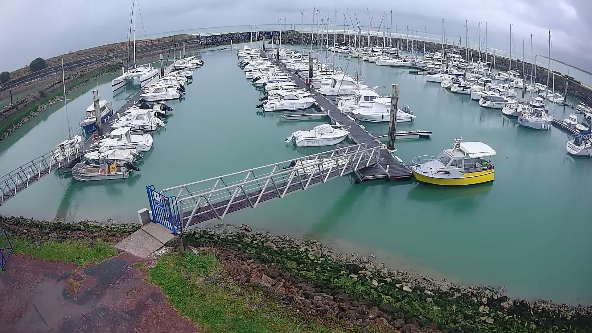 A marina filled with numerous boats and yachts on a cloudy day, with a metal walkway leading to a dock.