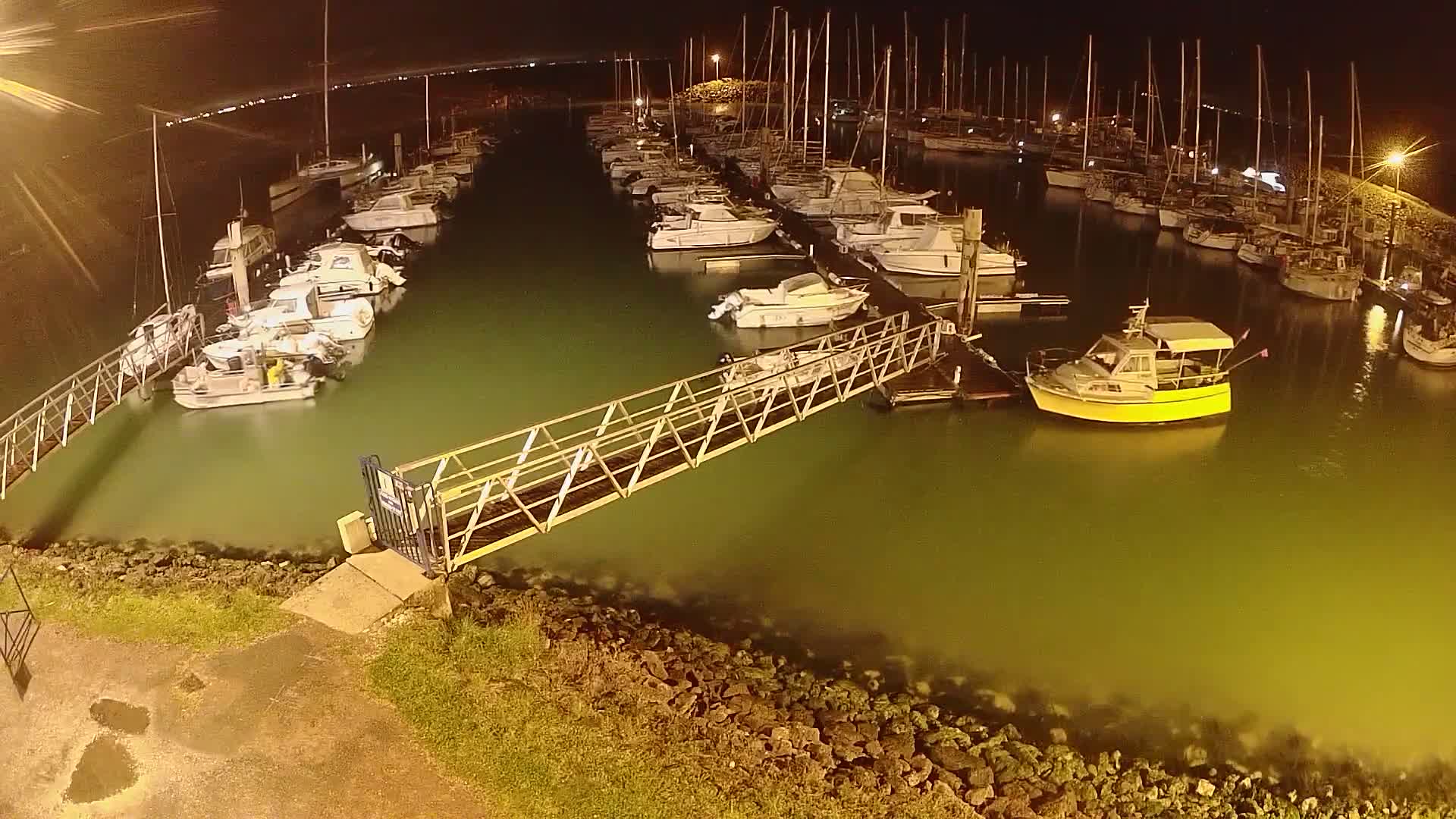 A marina at night, filled with numerous boats and yachts, is illuminated under artificial lights; a metal walkway connects the shore to a dock.