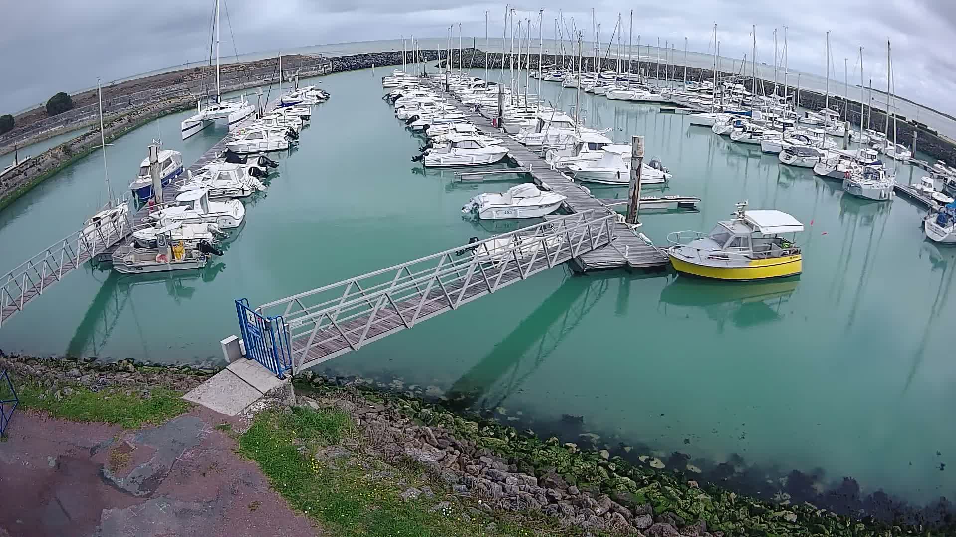 A marina filled with numerous sailboats and motorboats on a cloudy day, with a metal walkway leading to a dock.