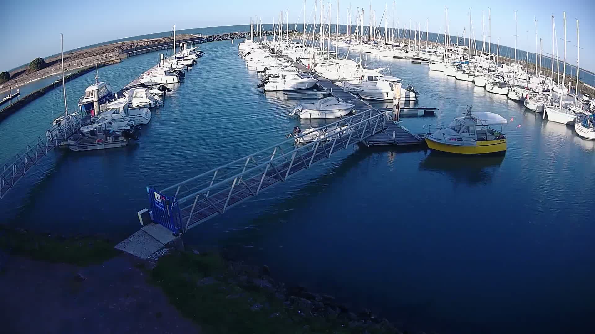 A marina filled with numerous boats and yachts on a sunny day, with a metal walkway leading to a dock.