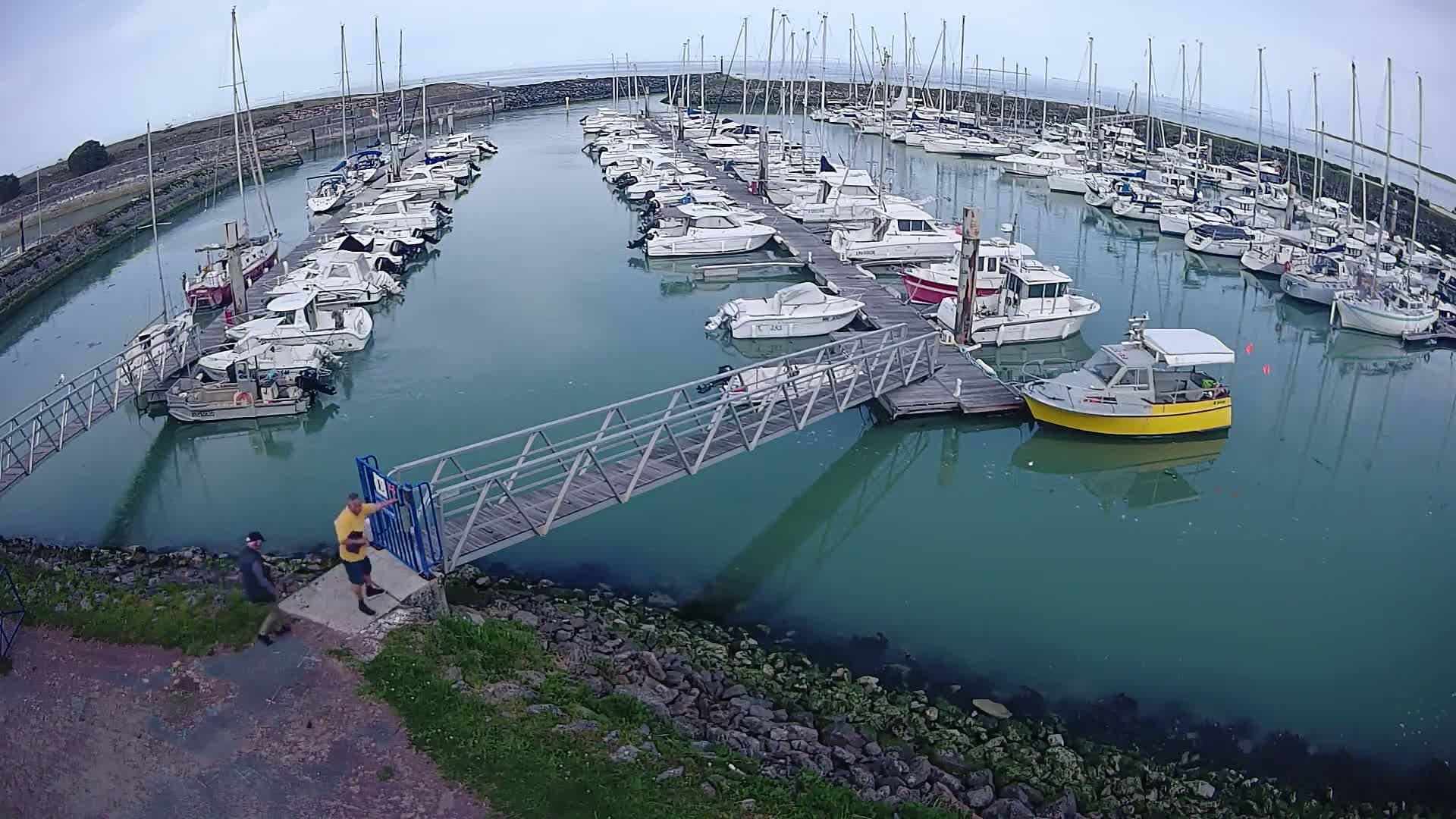 A marina filled with numerous sailboats and motorboats under a cloudy sky, with two people standing near a metal walkway leading to a dock.