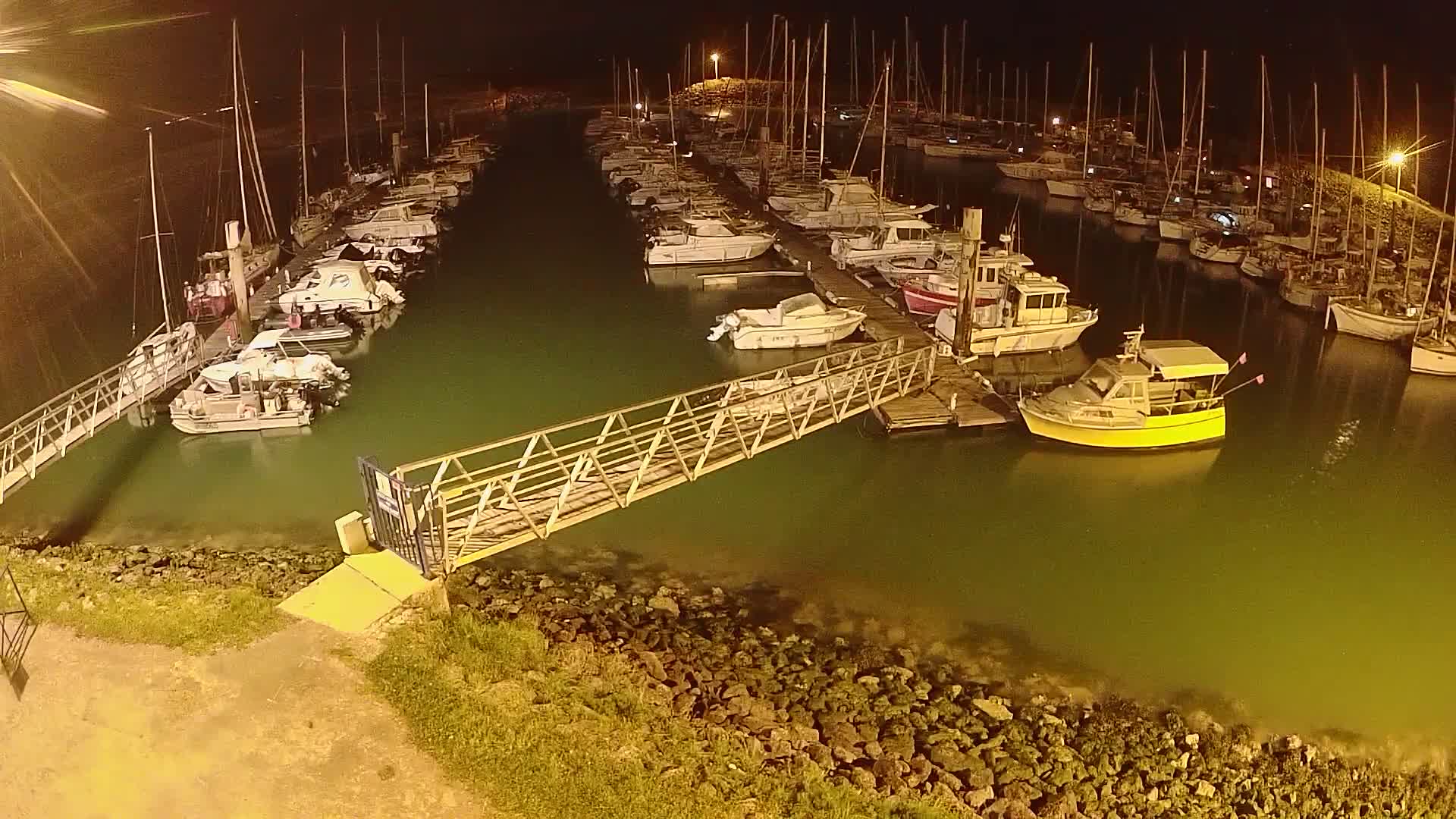 A marina at night, filled with numerous boats moored alongside docks, is illuminated by artificial lights, with a metal walkway extending from the shore to a central dock.