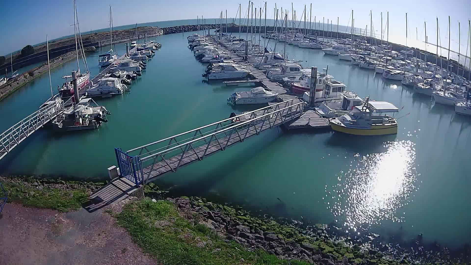 A marina filled with numerous boats and yachts on a sunny day, with a metal walkway leading to a dock.