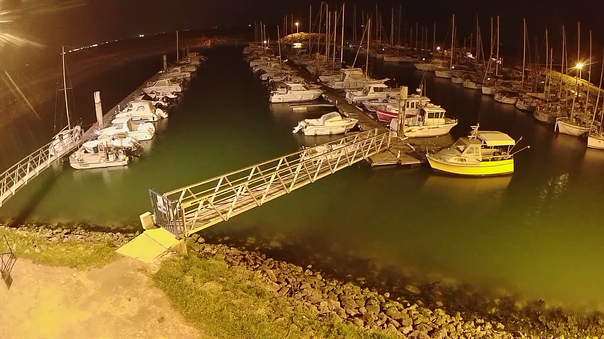 A marina at night, filled with numerous boats docked along piers, is connected to the shore by a metal walkway.