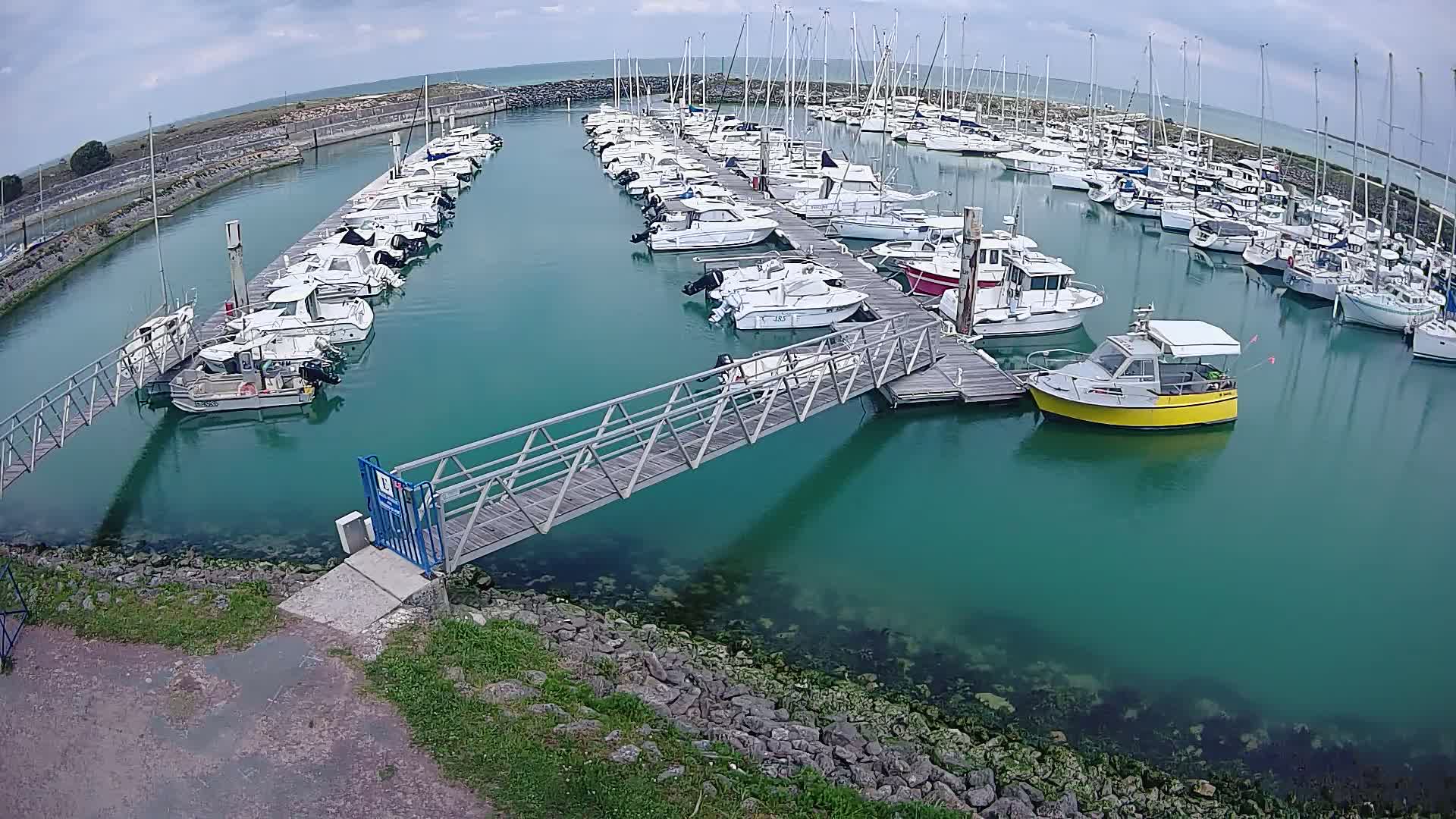 A marina with numerous boats docked at piers under a partly cloudy sky, connected by a metal walkway.