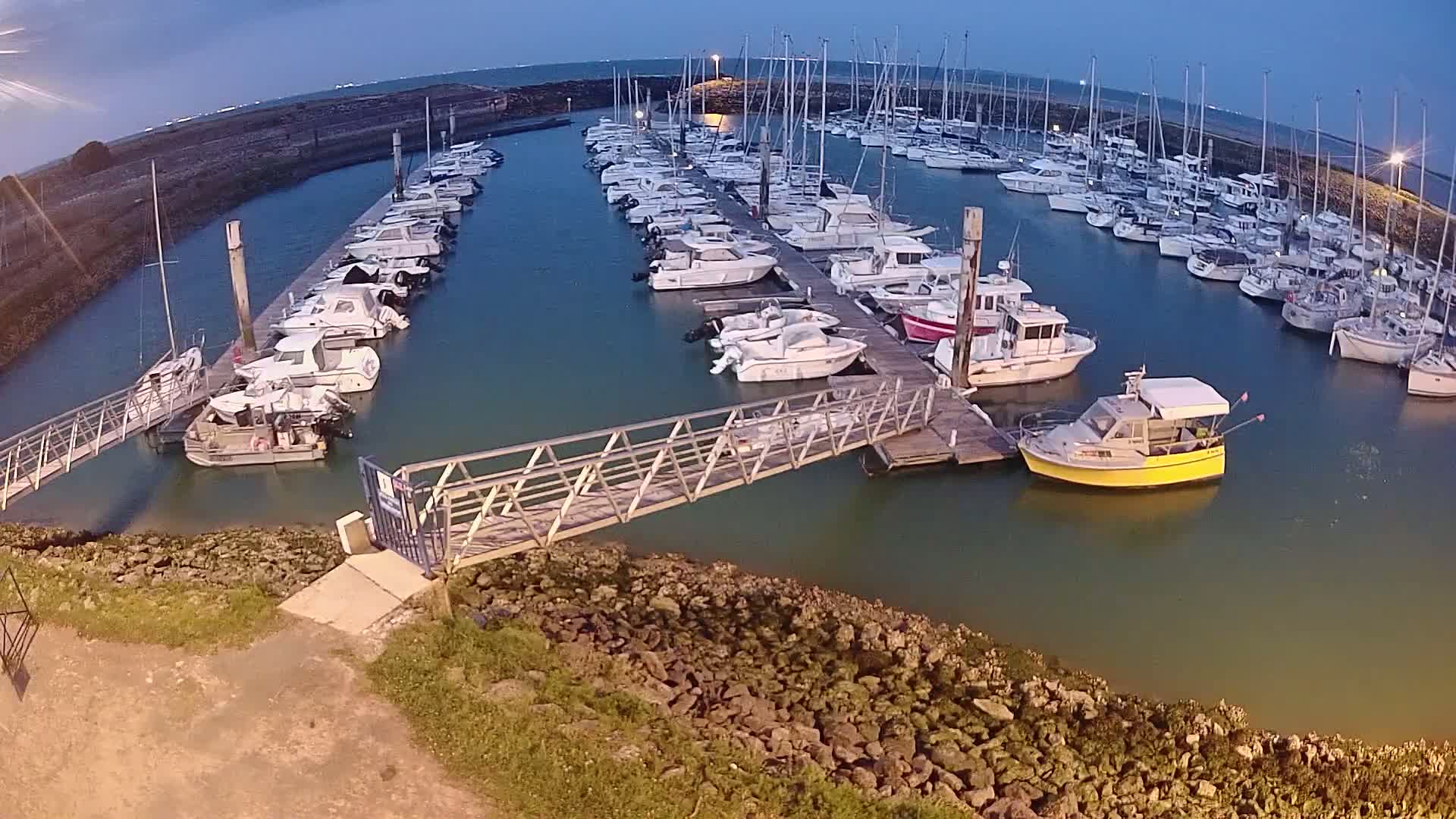 A marina at dusk filled with numerous boats docked along several piers, connected by a metal walkway.