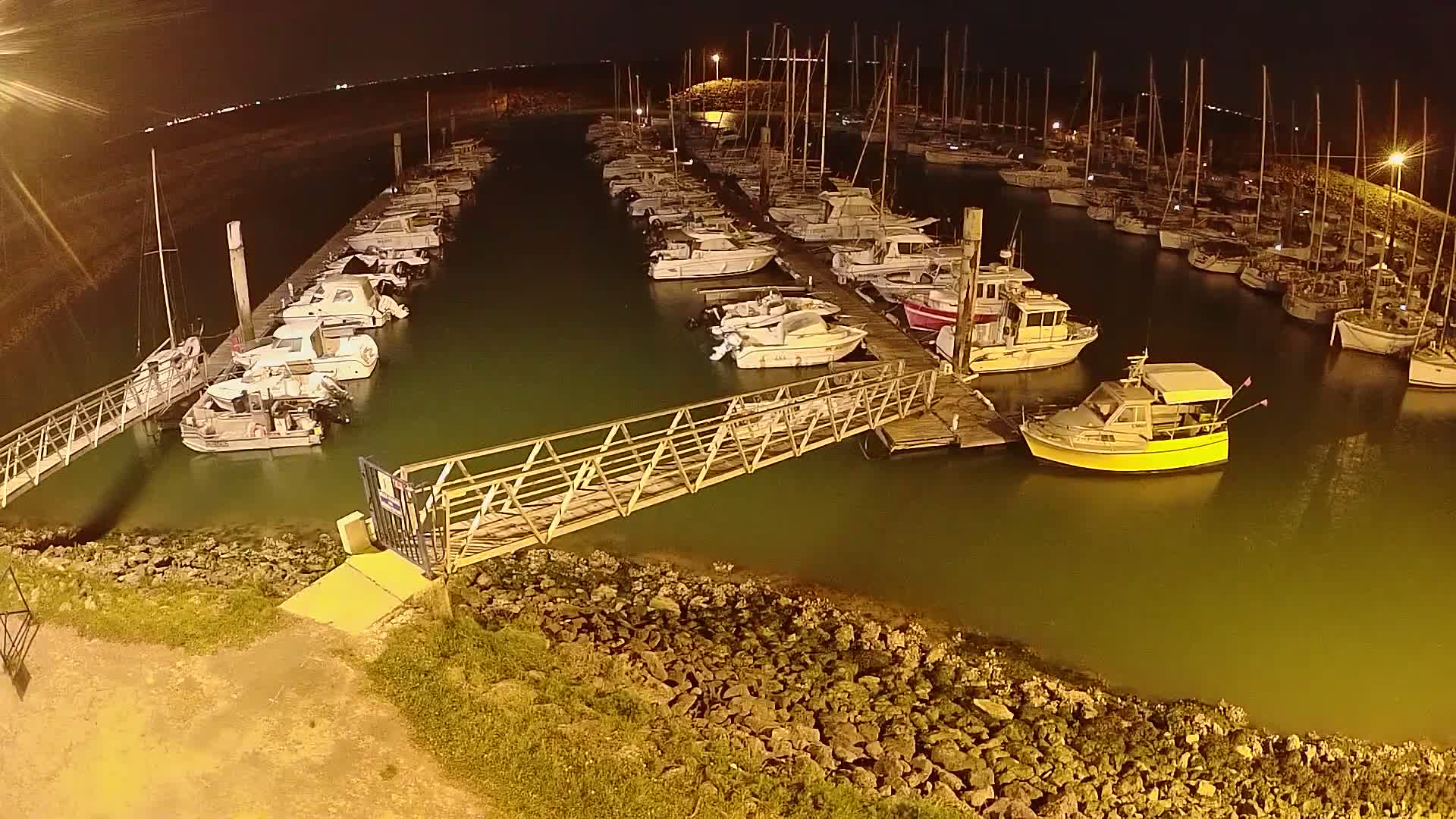 A marina at night, filled with numerous boats docked along piers, illuminated by artificial lights, is seen from an elevated vantage point.