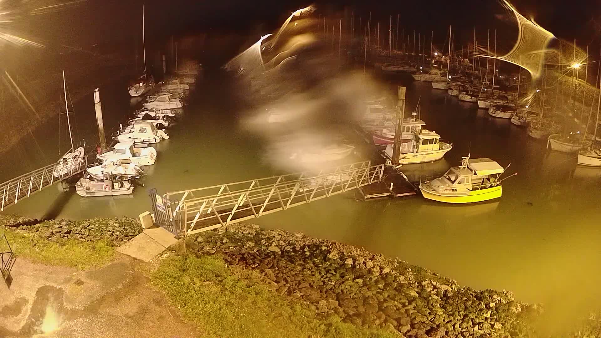 A marina at night, filled with numerous boats moored along docks, is illuminated by artificial light, with a metal walkway leading to a section of docked vessels.